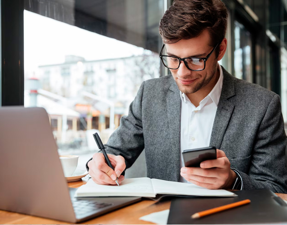 Profissional organizando informações em um caderno, ao lado de um notebook e com um celular na mão.
